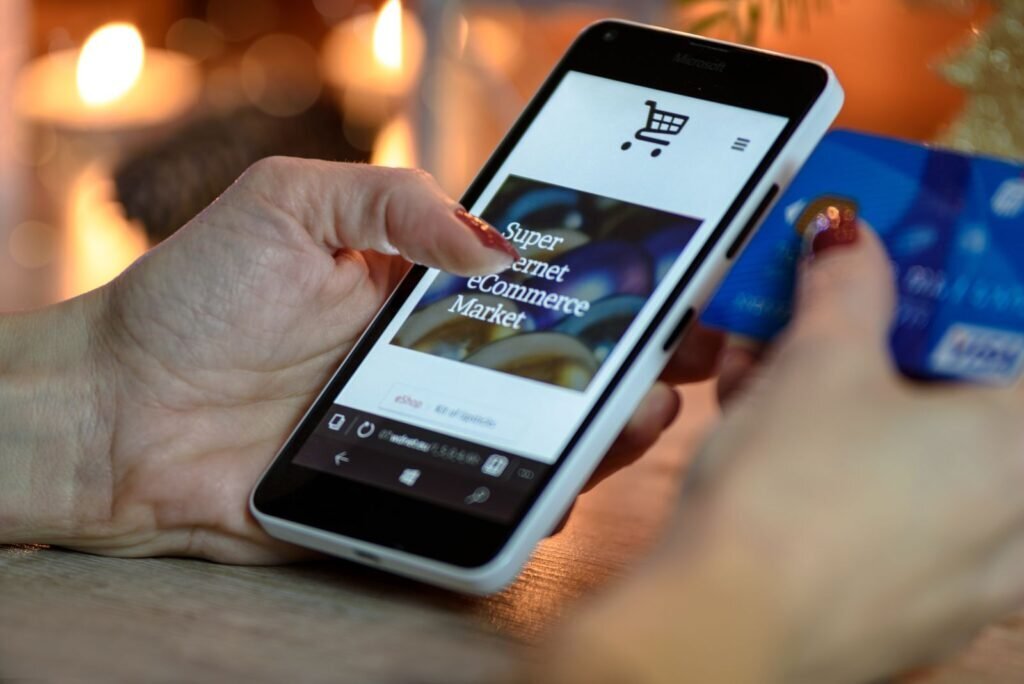 Hands holding a smartphone displaying an e-commerce site with a shopping cart, and a credit card. Represents mobile shopping and Google Shopping ad conversions.