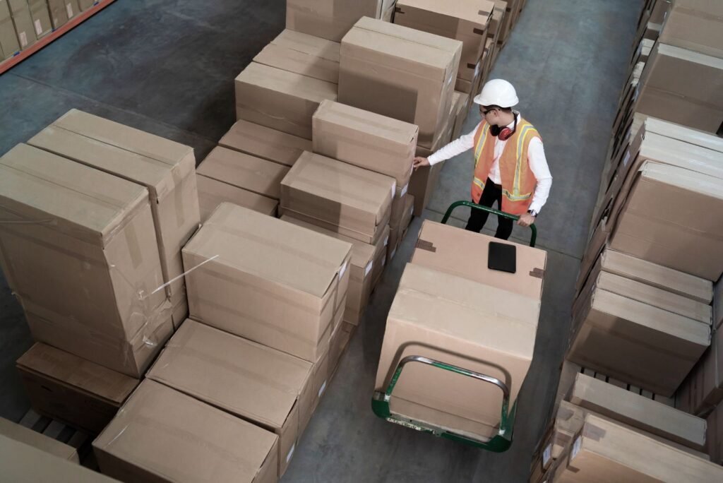 Thai logistics worker in a warehouse moving supply chain boxes with a hand truck, emphasizing digital inventory management.