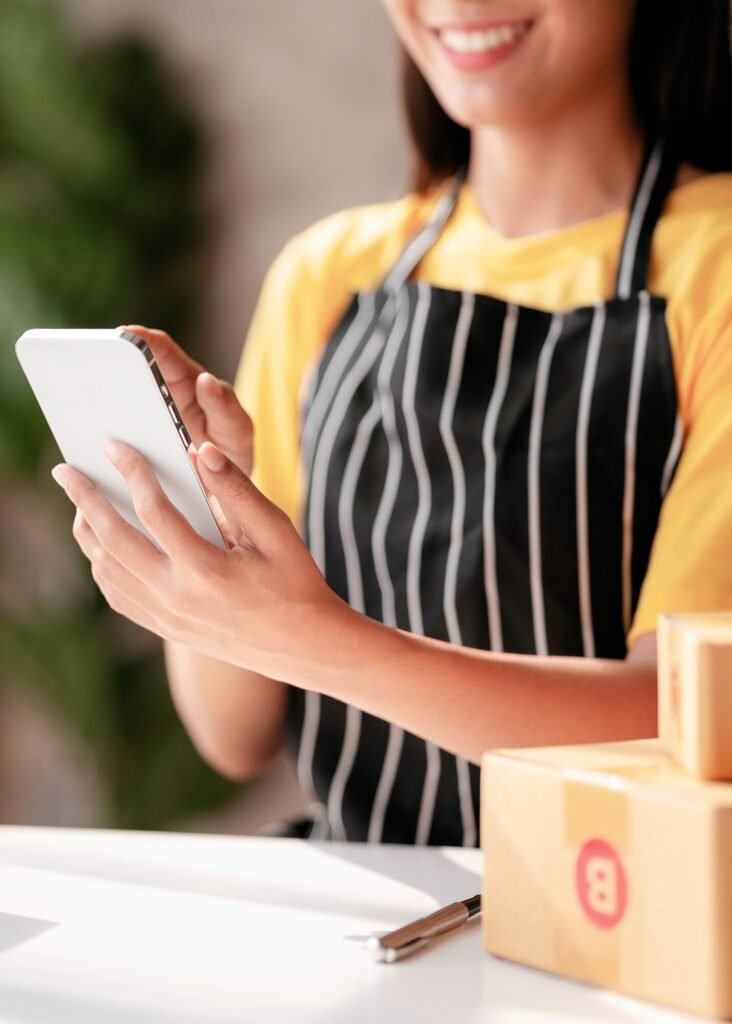 Smiling e-commerce business owner in a striped apron uses a smartphone, managing online orders and driving sales. Shipping boxes are ready.