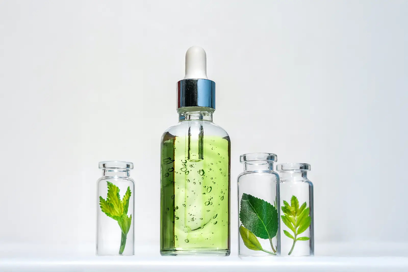 Goodganic organic serum bottle with green liquid, surrounded by vials of fresh leaves, showcasing natural ingredients.