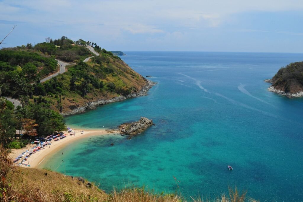 Aerial view of a beautiful Phuket bay with clear turquoise water, a sandy beach, and a winding road on a lush hillside.