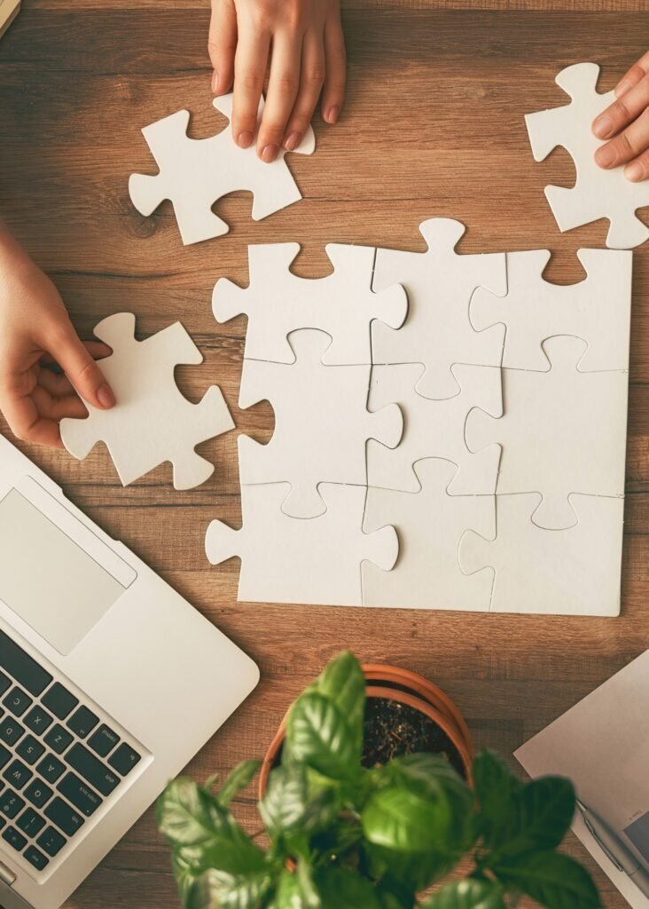 Multiple hands building a white jigsaw puzzle on a desk with a laptop, representing collaborative CPAS strategy for sales.