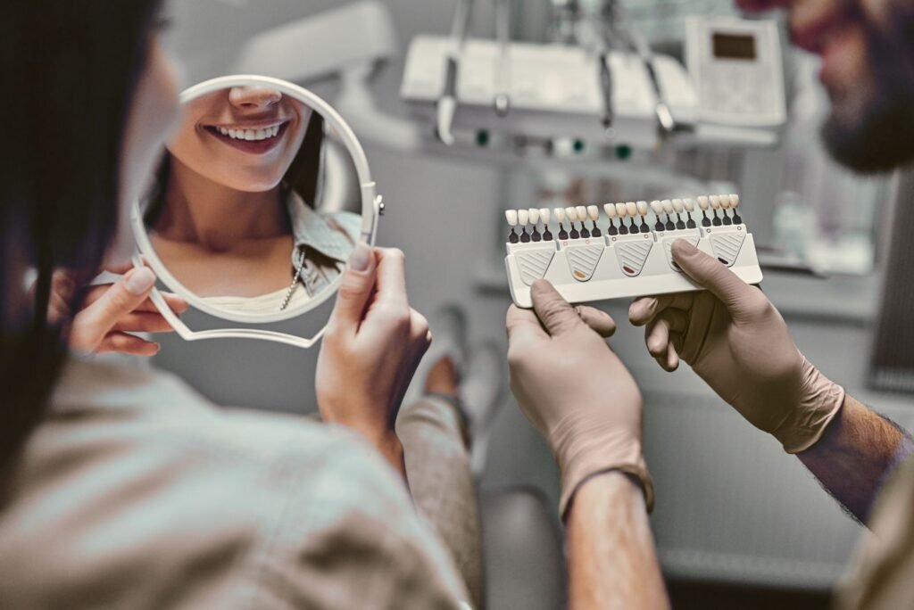 Happy patient views her bright smile in a mirror while a dentist holds a tooth shade guide for cosmetic dental services.