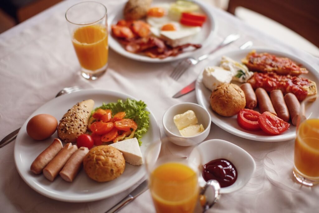Abundant breakfast buffet with fried eggs, crispy bacon, sausages, fresh vegetables, cheese, warm bread, and orange juice.