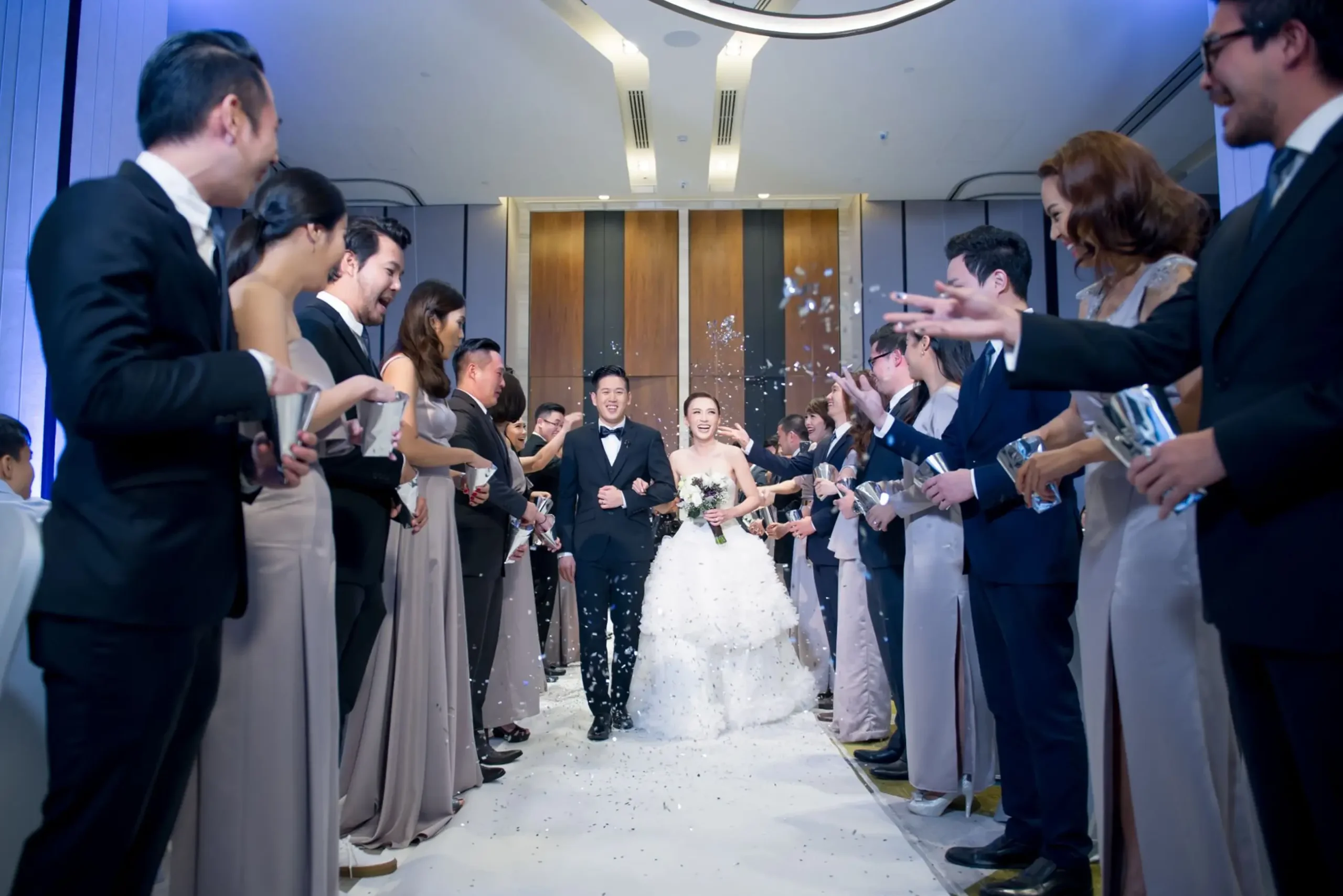 Smiling Thai wedding couple and guests celebrating with confetti in a modern hotel ballroom, showcasing hospitality.