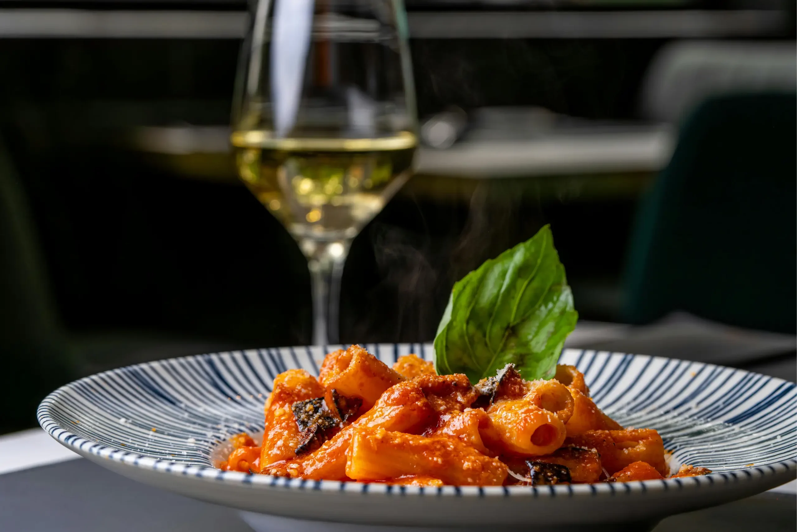 Steaming rigatoni pasta with rich tomato sauce, fresh basil, and grated cheese, paired with white wine, for hospitality marketing.