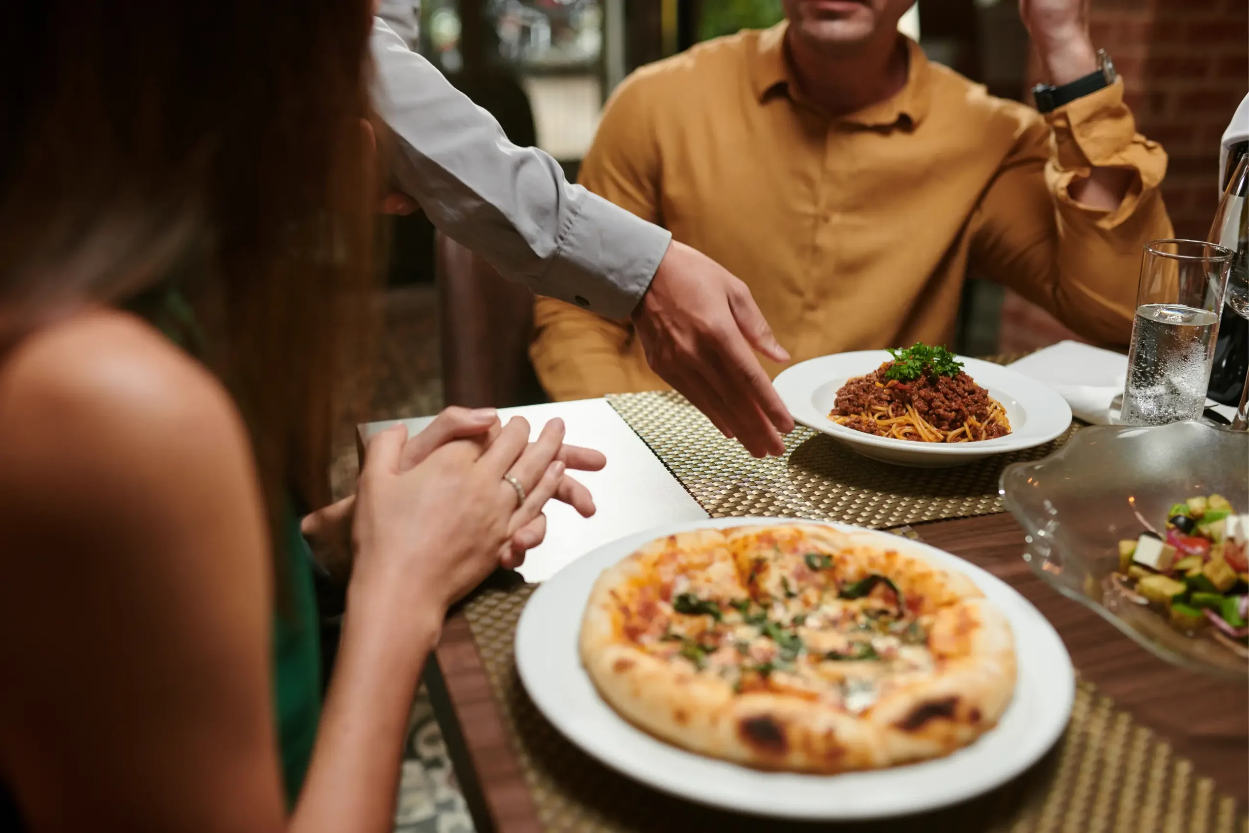 Server places spaghetti for guests at a restaurant table with pizza, reflecting excellent hospitality.